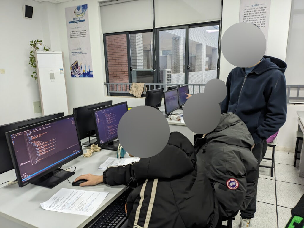 Three students in a computer classroom, with two students working on their respective computers and one student standing behind them.