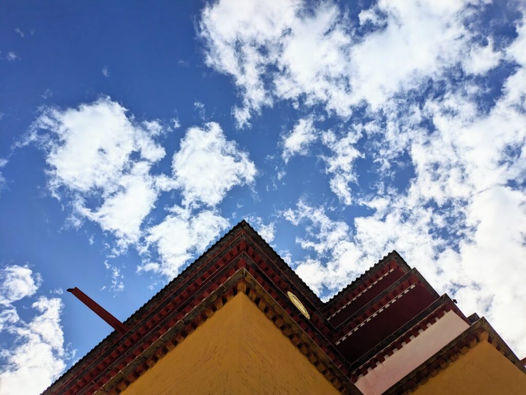 From a bottom-top view, blue sky with clouds along with the corner of a Tibetan building with a yellow wall on the bottom.