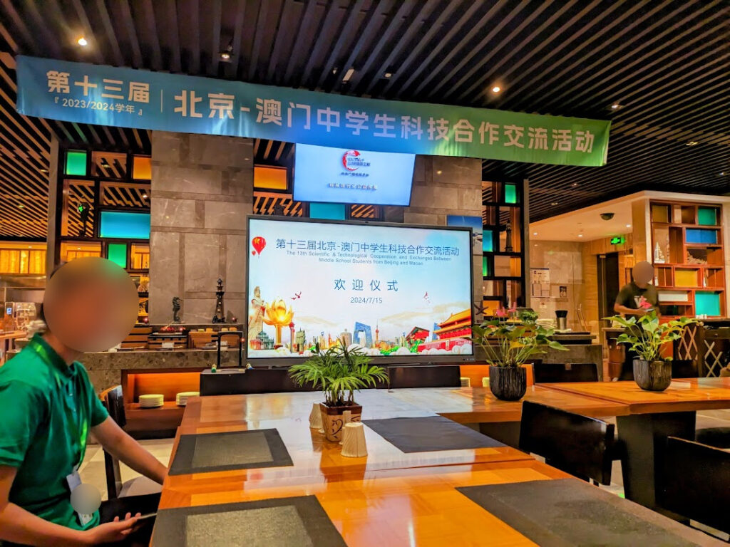 In a hotel room at night, a banner hanging from the ceiling reading "13th Beijing Macao High School Students Sci-Tech Coop and Exchange", along with a screen showing "Welcome".