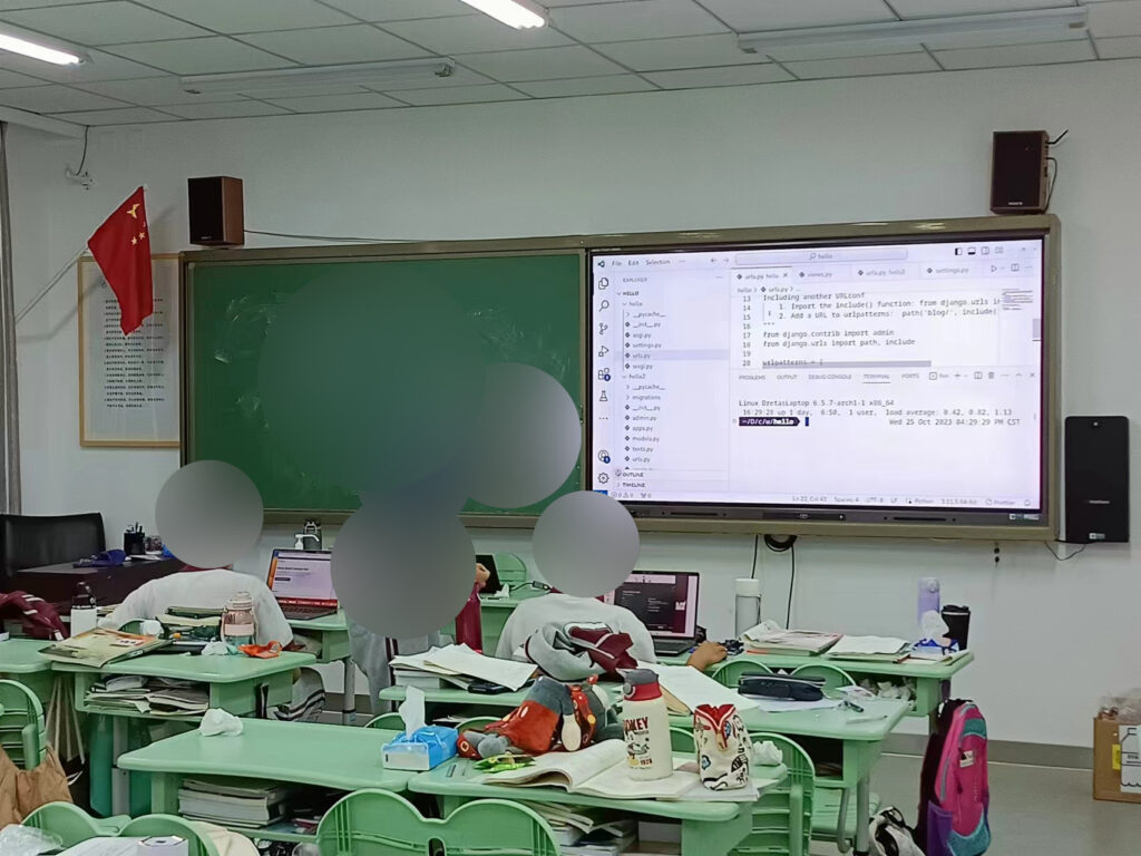 3 students in a classroom with a blackboard and a screen showing web development software up front. One student is tutoring another.
