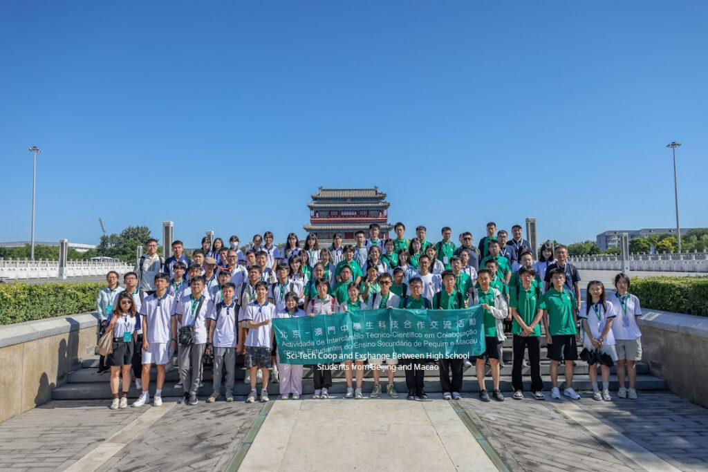 A group photo taken under a clear blue sky before Beijing's Yongdingmen, with the students in the front holding a banner reading "Sci-Tech Coop and Exchange Between High School sStudents of Beijing and Macao".