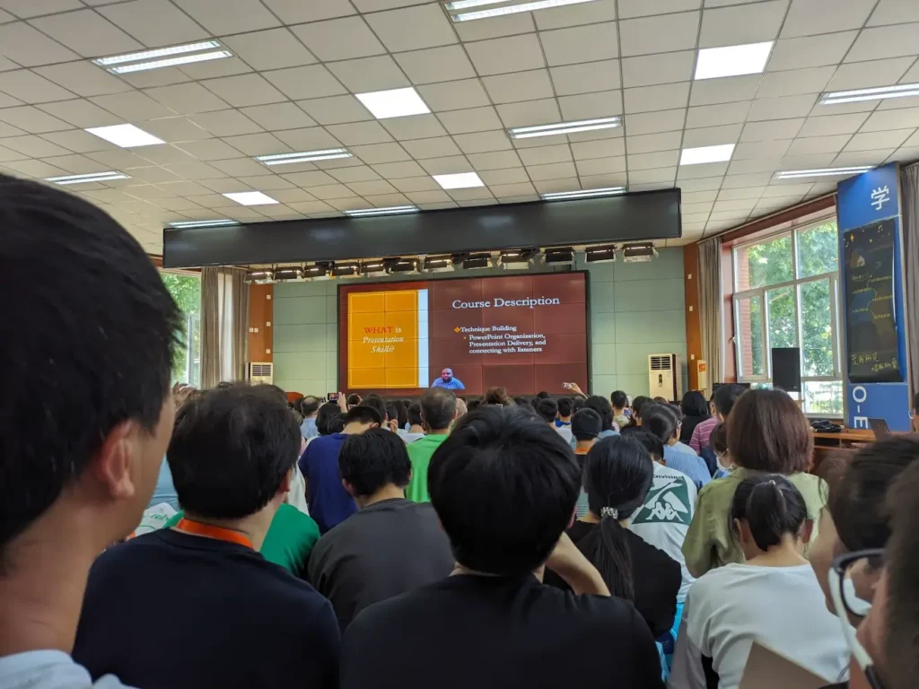 A huge crowd sitting in an assembly hall. A teacher is presenting the presentation course syllabus.