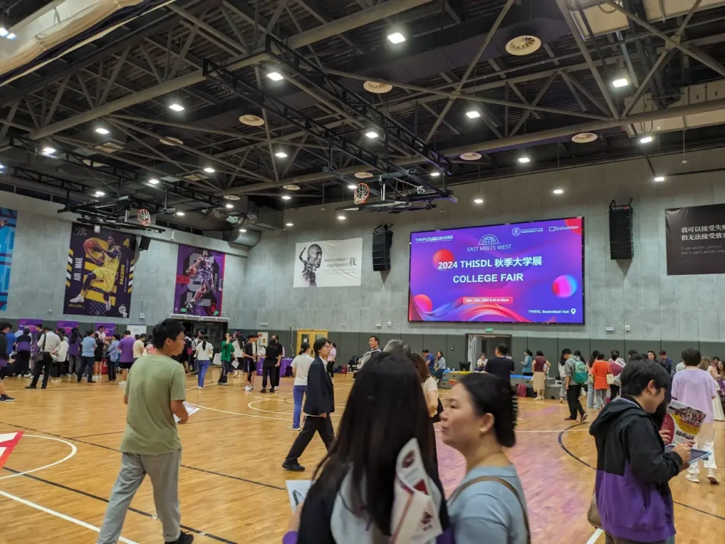 A gym in THISDL, the private international school that's hosting the college fair. There's a screen with the text "2024 THISDL Fall College Fair" on it, and many people inside the gym.