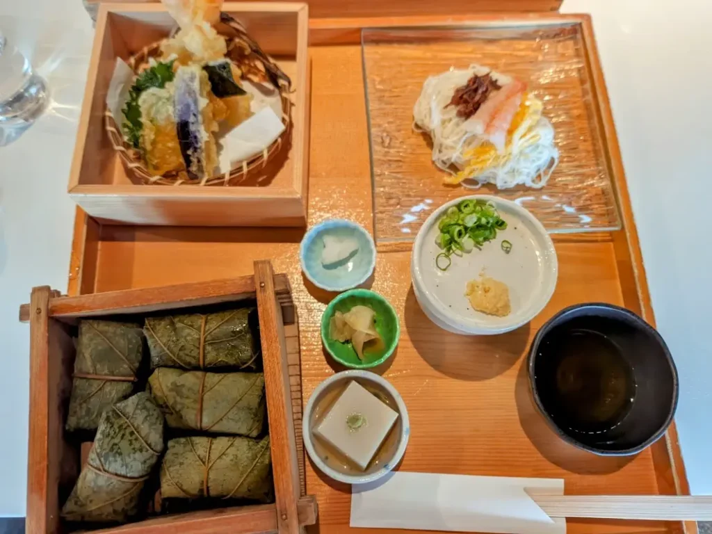 A wooden plate with shrimp udon, a bowl of miso soup, tempura, and five persimmon-leaves sushis on it.