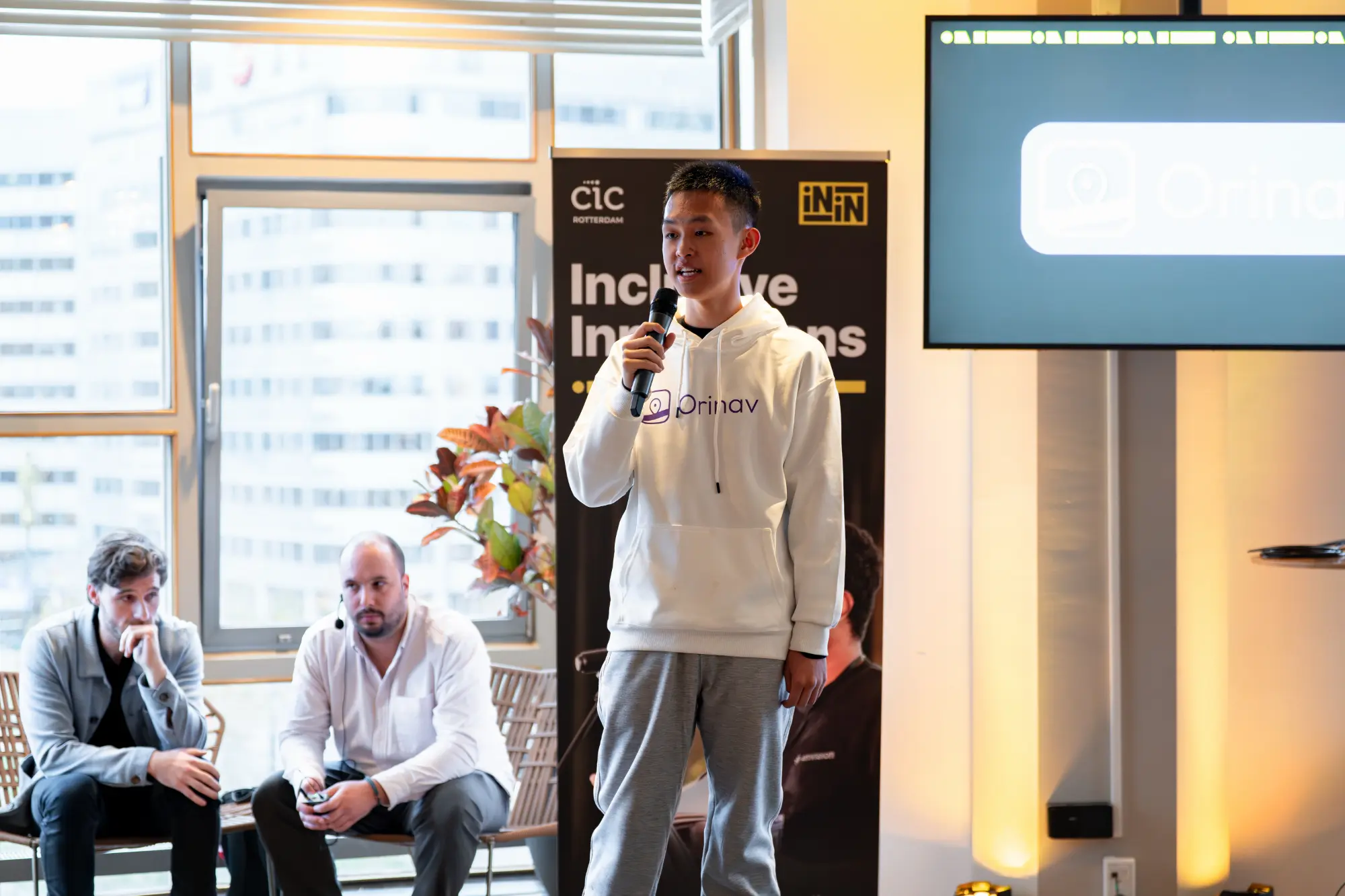 Lin wearing an Orinav hoodie, holding a microphone speaking at Inclusive Innovations.