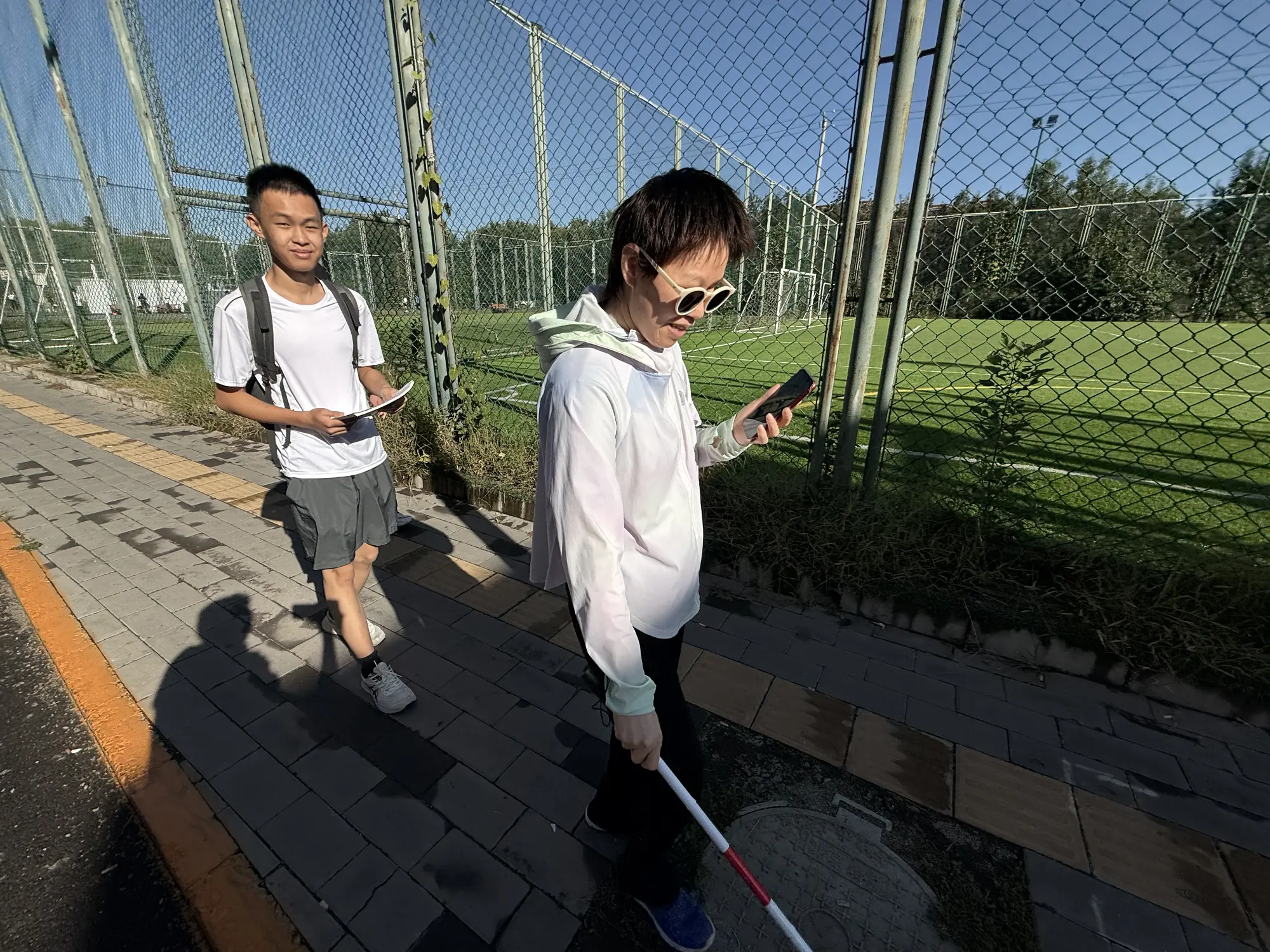 A woman with blindness using a white cane and Orinav to navigate. Lin is behind her, recording.